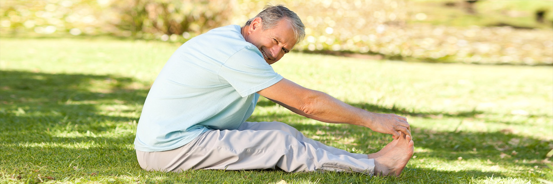 Mature man sitting on grass in park touching toes, practicing Yoga